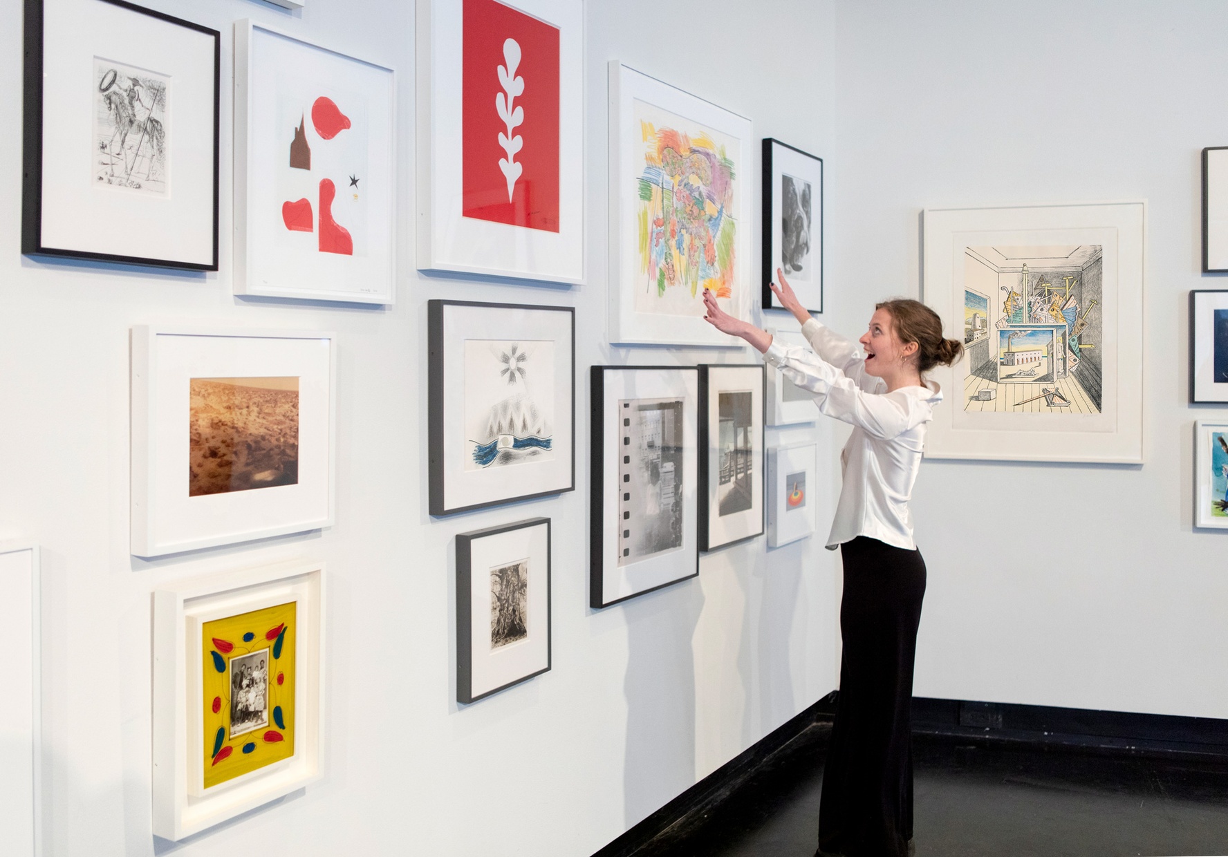 A student stands smiling in front of a wall of framed artwork with her hands reached out towards a large print by Carroll Dunham.