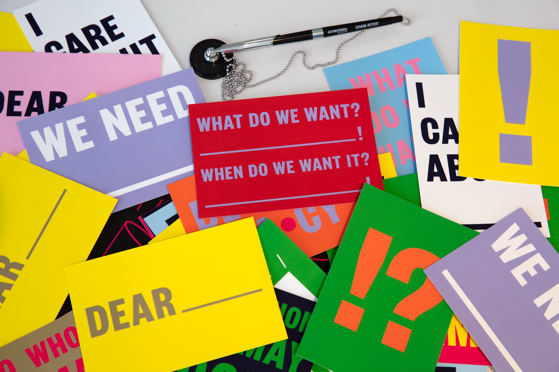 Various colorful postcards with text like "Dear___", "We Need ____", and "What do we want? _____! When do we want it? ______!" spread out on a table.