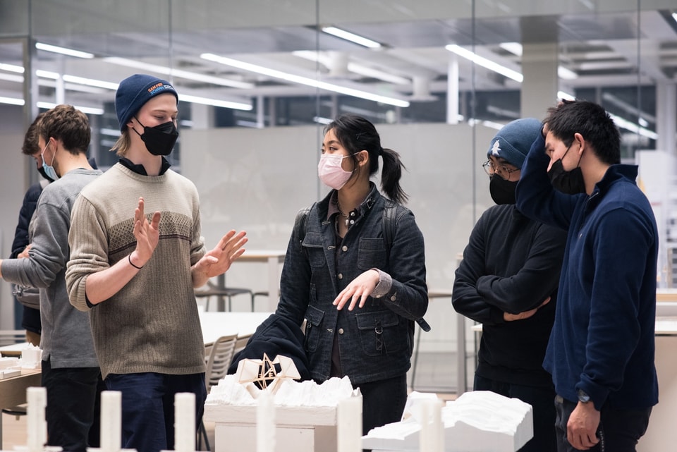 Three people animatedly discuss a white architectural building model sitting on a low table beside them.