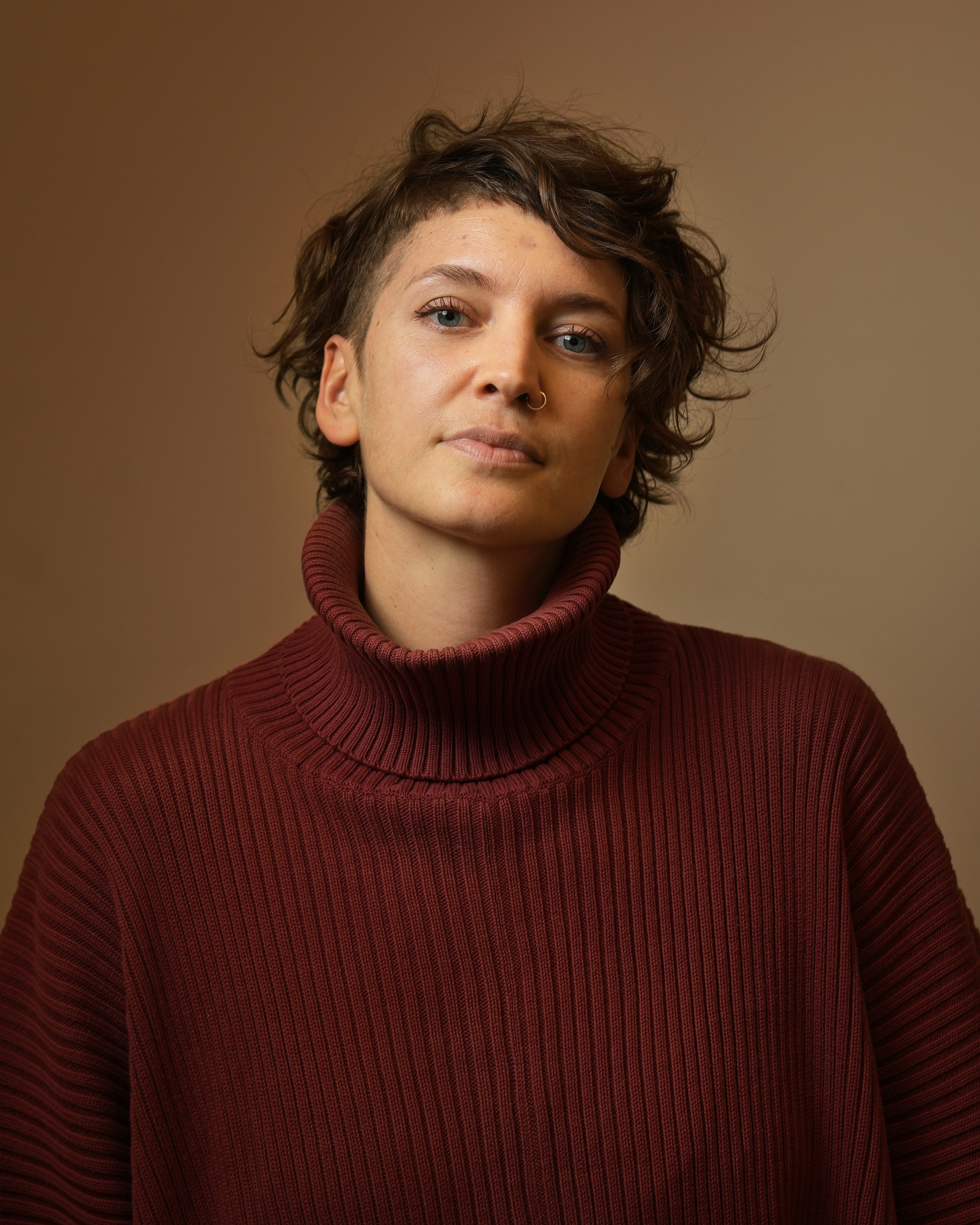 A portrait of Zoe Rabinowitz against a neutral brown background. Zoe wears a wine-red turtleneck sweater and tilts her head back slightly, looking directly at us. 