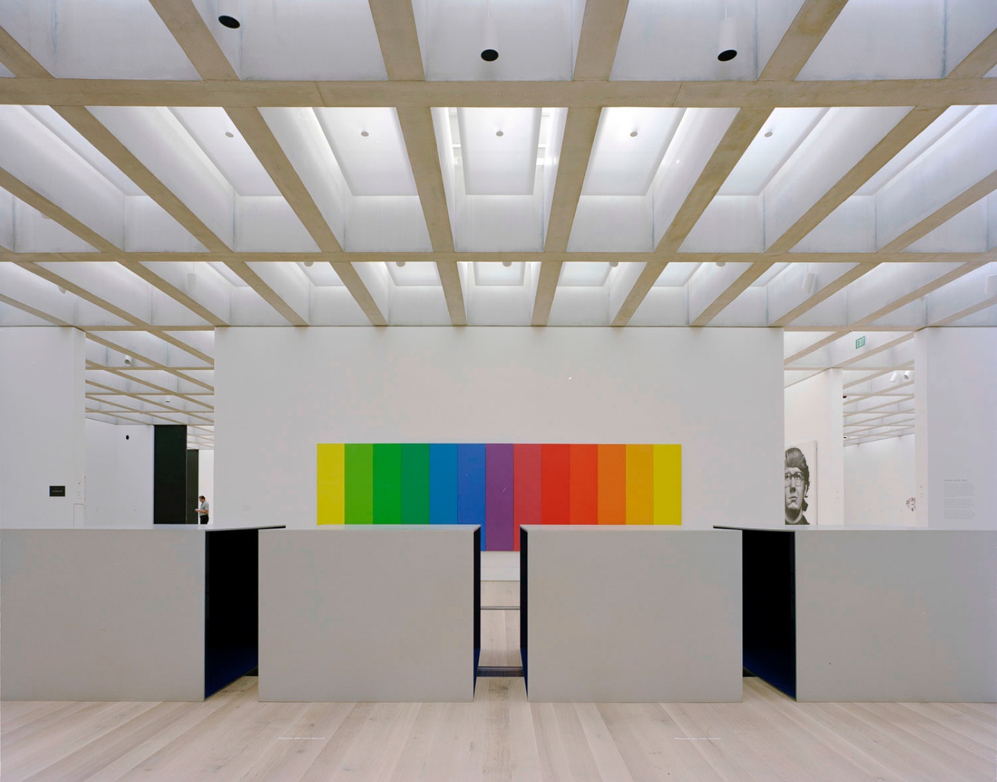 Interior gallery space featuring a high ceiling with perpendicular beams. A paneled, rainbow-colored artwork is displayed on the back wall, with four square pedestals in the foreground.