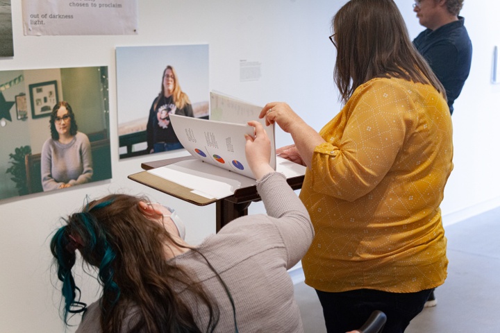 Two gallery visitors flip through a three-ring binder placed on a podium. The displayed page shows three pie charts with data. The podium faces a wall with portrait photographs of two young white women in casual clothes.