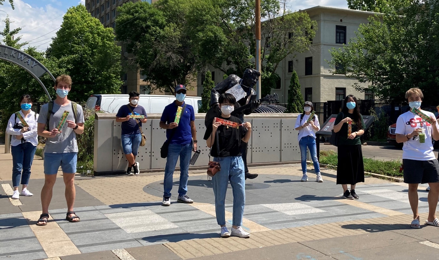 Eight students stand in three rows, spaced at least 6 feet apart from each other and wearing protective face masks; they are also holding rulers to measure social distance. They are standing outside in University City, Missouri.