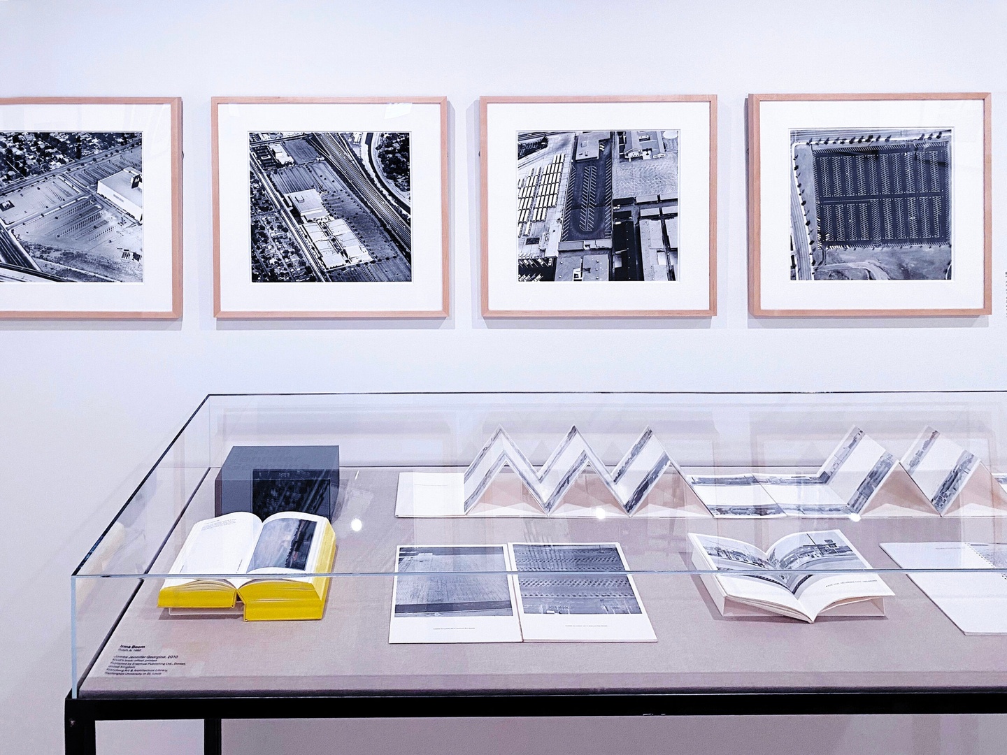 In a museum exhibition, four framed black and white images are hung on the wall. Below them is a table with a glass case. Inside, several books containing black and white images lay open.
