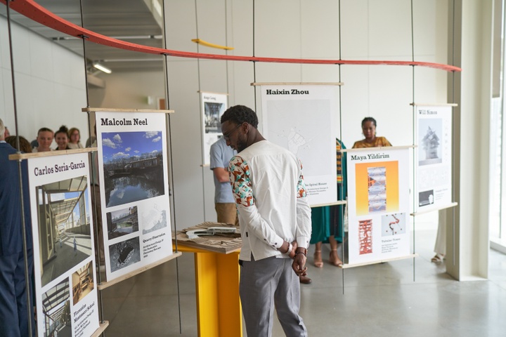 Student views an architectural exhibition composed of poster panels suspended at varying heights from a red, curving beam. Each poster has a student's name and three images of their work. Physical models are placed on bright yellow and red stands around the space.
