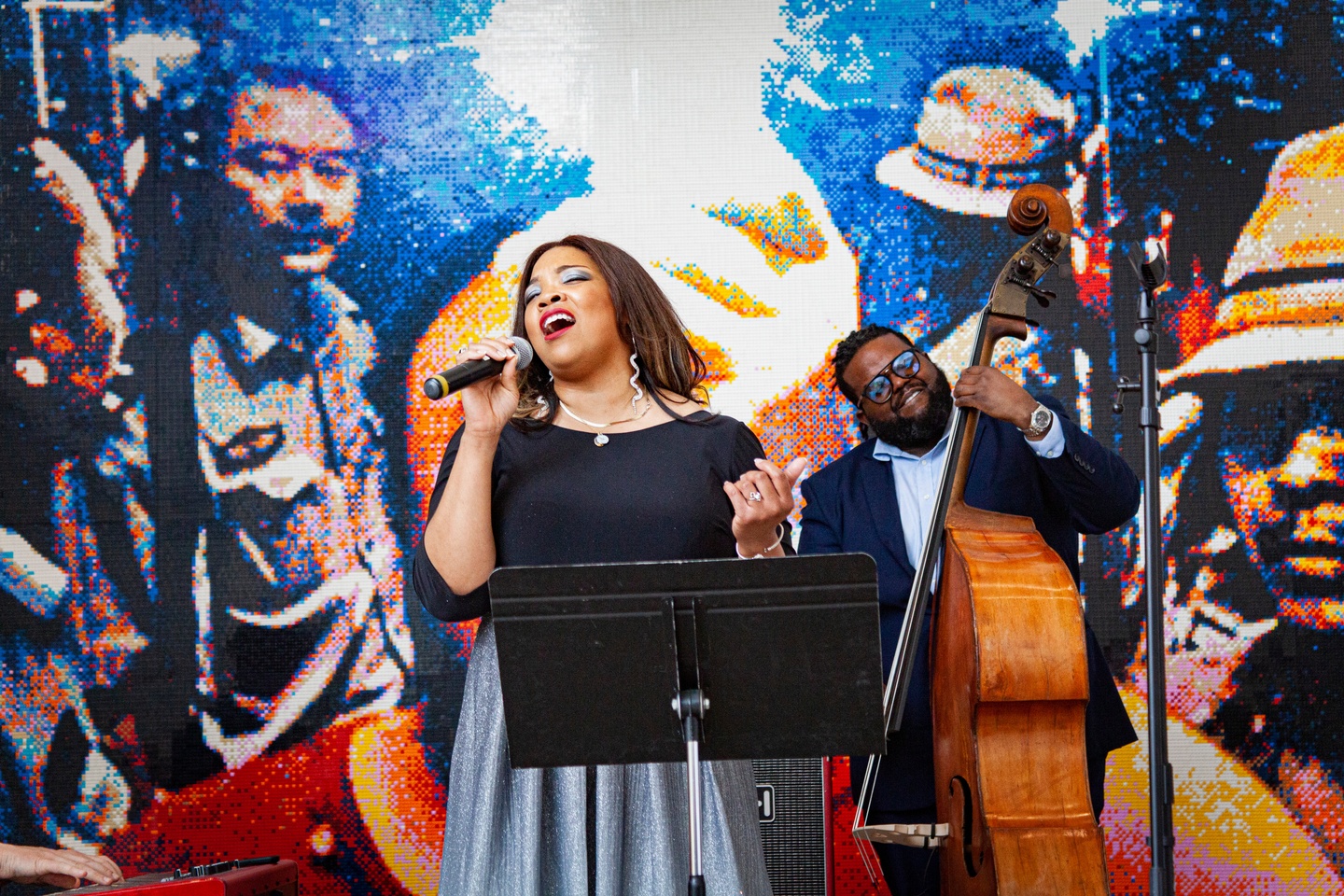 A woman singing into a microphone and a man playing an upright bass stand before a colorful artwork that includes people's faces