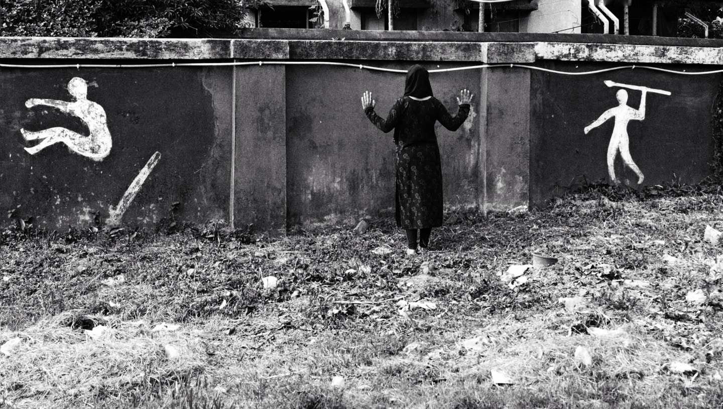 black and white photo of a woman standing and facing a wall with her hand up covered head to toe. Painted on the left side is a silhouette of a person jumping and on the right side is a silhouette of a person with a spear