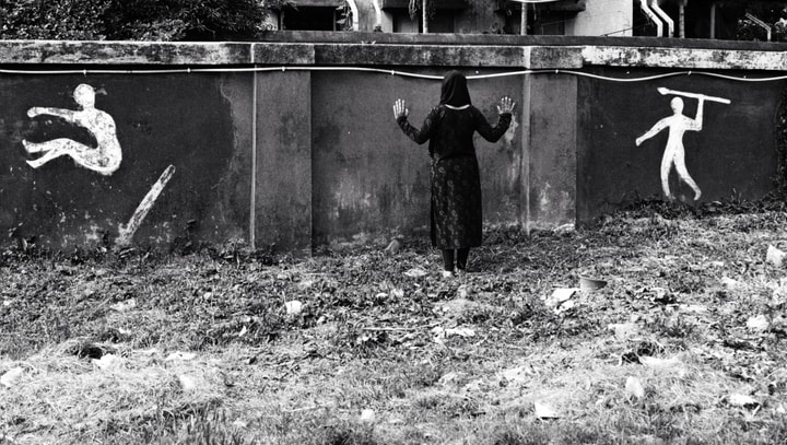 black and white photo of a woman standing and facing a wall with her hand up covered head to toe. Painted on the left side is a silhouette of a person jumping and on the right side is a silhouette of a person with a spear