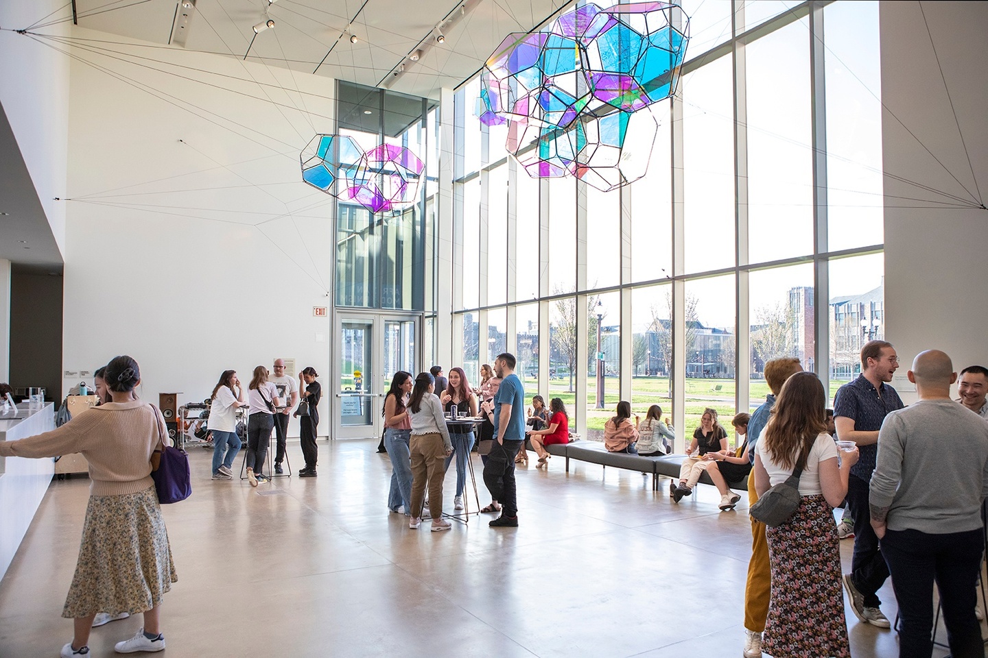 A photograph of people standing in groups and sitting in a gallery space with a colorful artwork hanging above