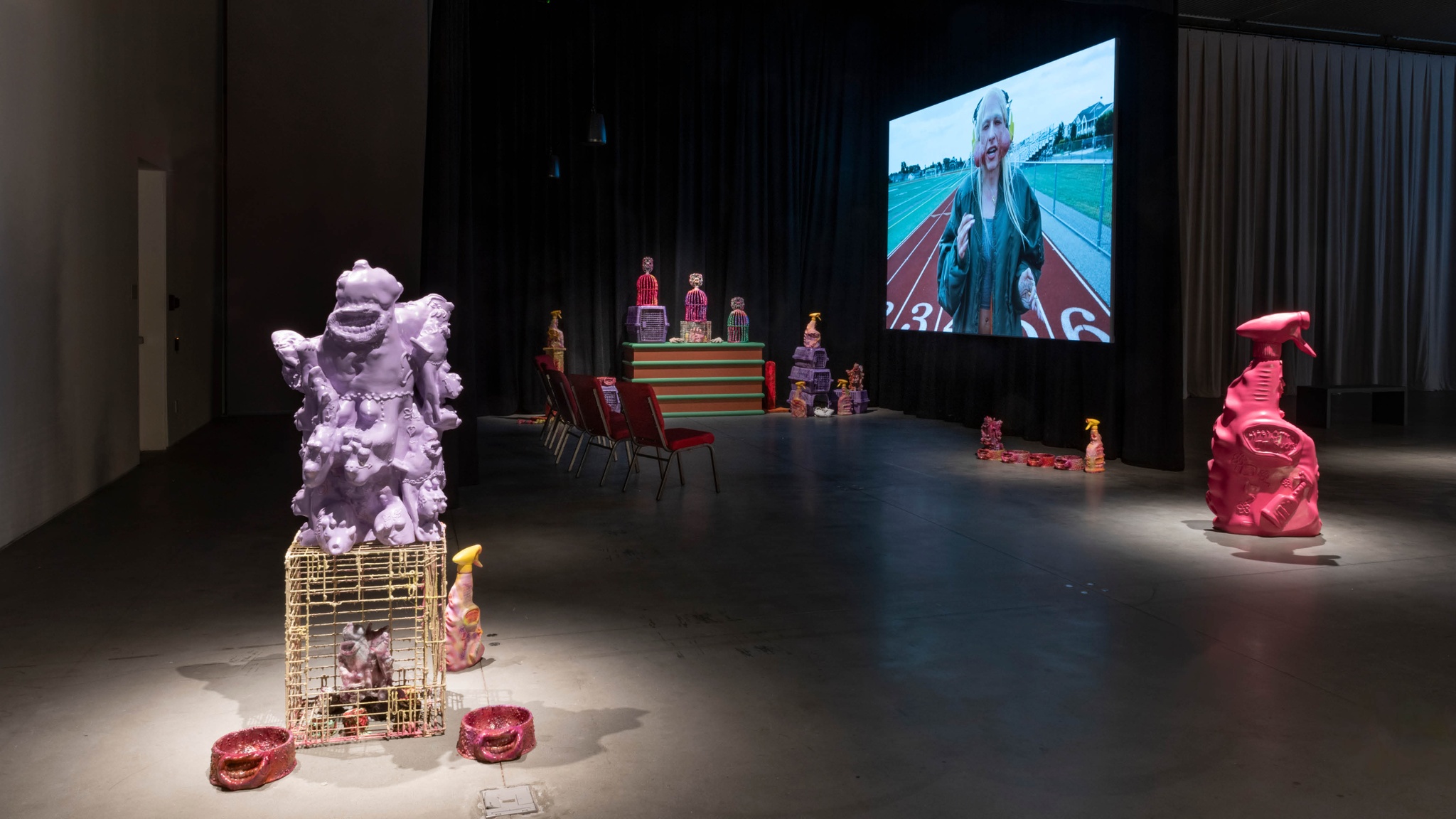 An installation of sculptural objects and a film in a gallery with low light. The sculptural objects are oversized versions of pet products, like cages, dog bowls, and cleaning supplies. They are cast in fluorescent colors. In the foreground is a large pink spray bottle sculpture, about four feet tall. In the background, the film projected on a screen shows the artist Jake Brush in character with a long blond wig and prosthetic jowls protruding from his cheeks while he walks on a high school track.
