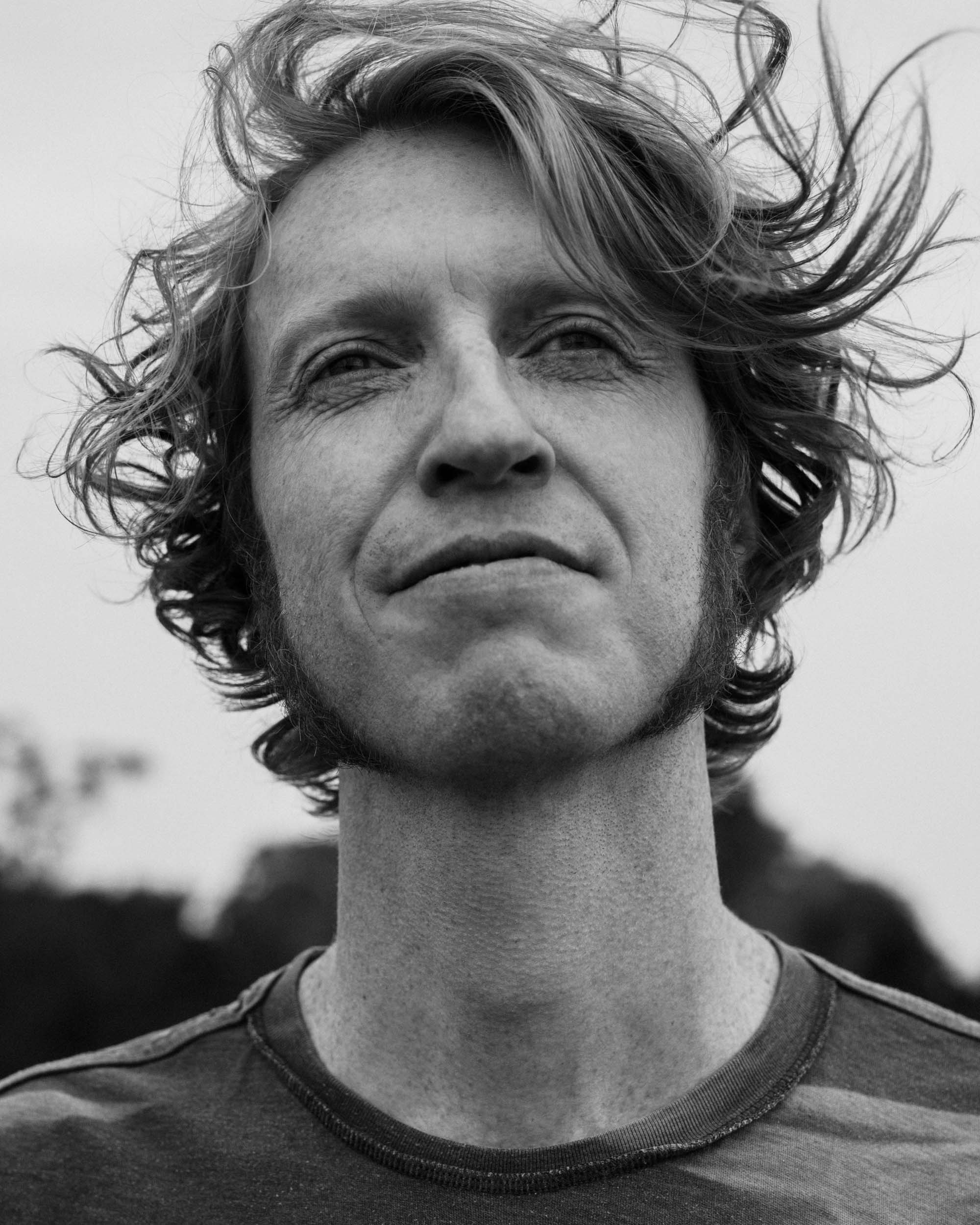 A white man seen from below, gazing off into the distance. He has unkempt hair that swoops down across his forehead in this black and white photo. 