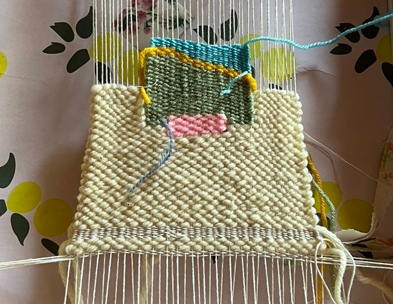 Closeup of a loom with beige, pink, green and blue thread woven through it