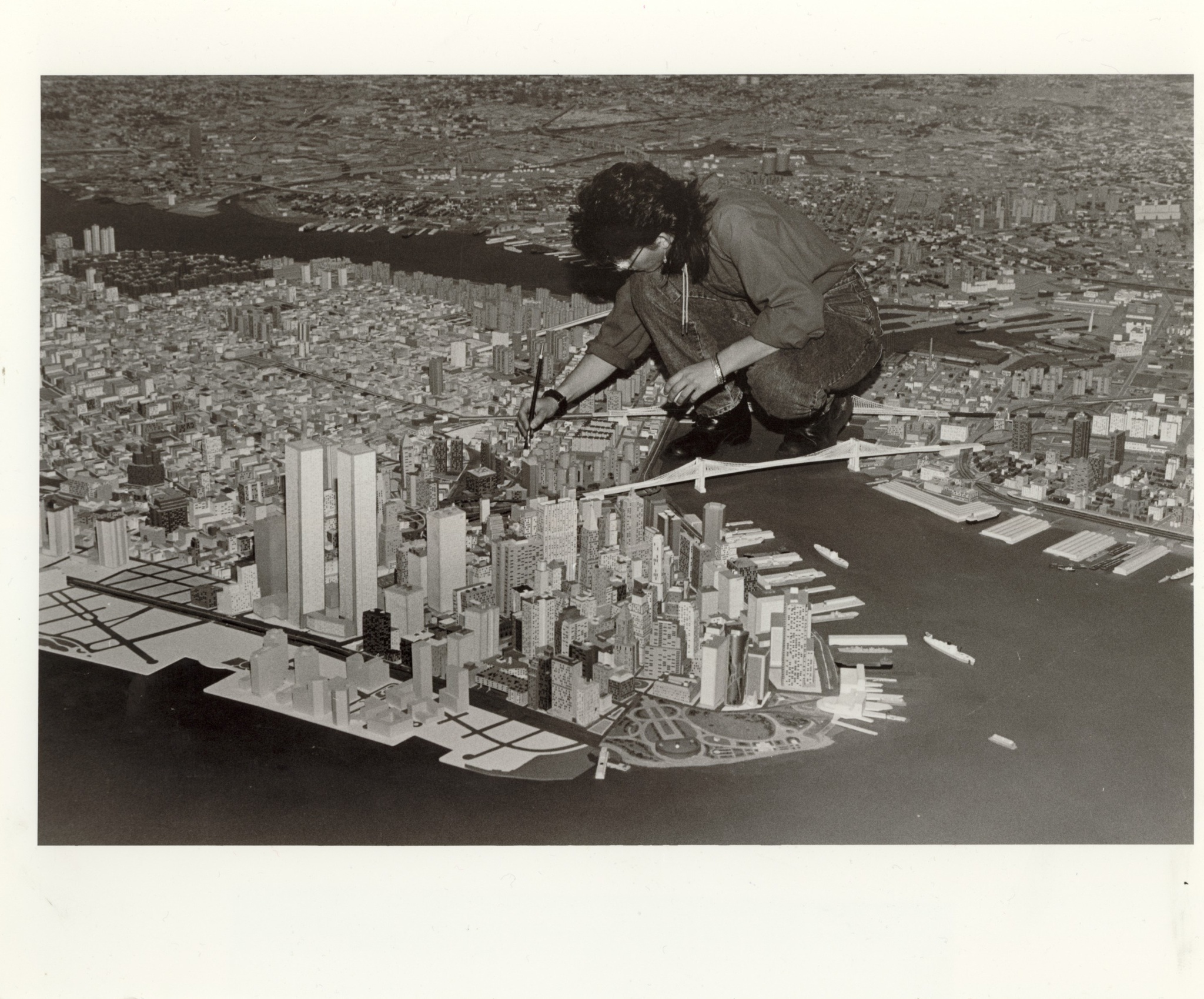 A person crouches atop the section of the Panorama of the City of New York that represents the East River, hunched over making repairs to the model buildings that make up Manhattan.