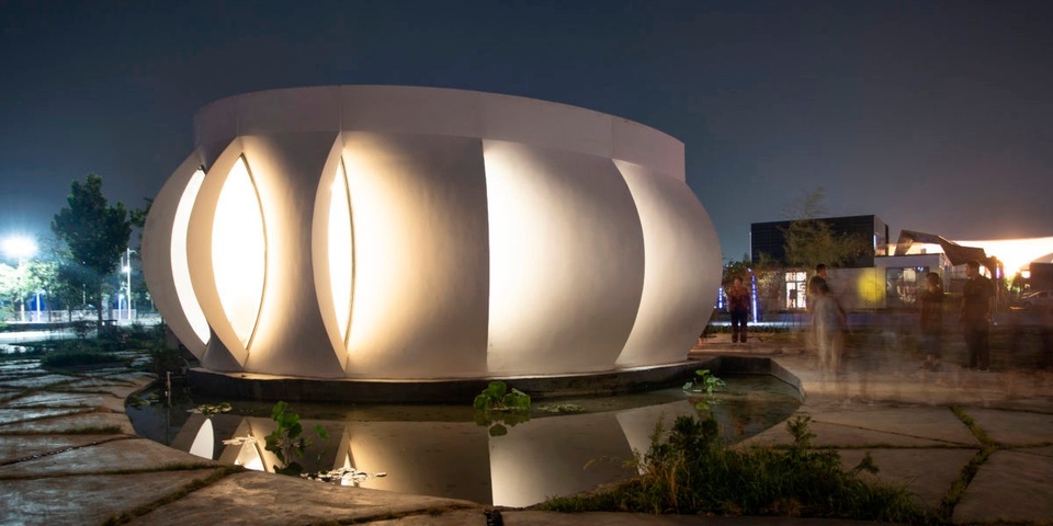 Photo of the exterior facade of a single-story home lit up at night. The exterior is composed of curved, overlapping panels, arrayed around a central axis like a blossoming flower.
