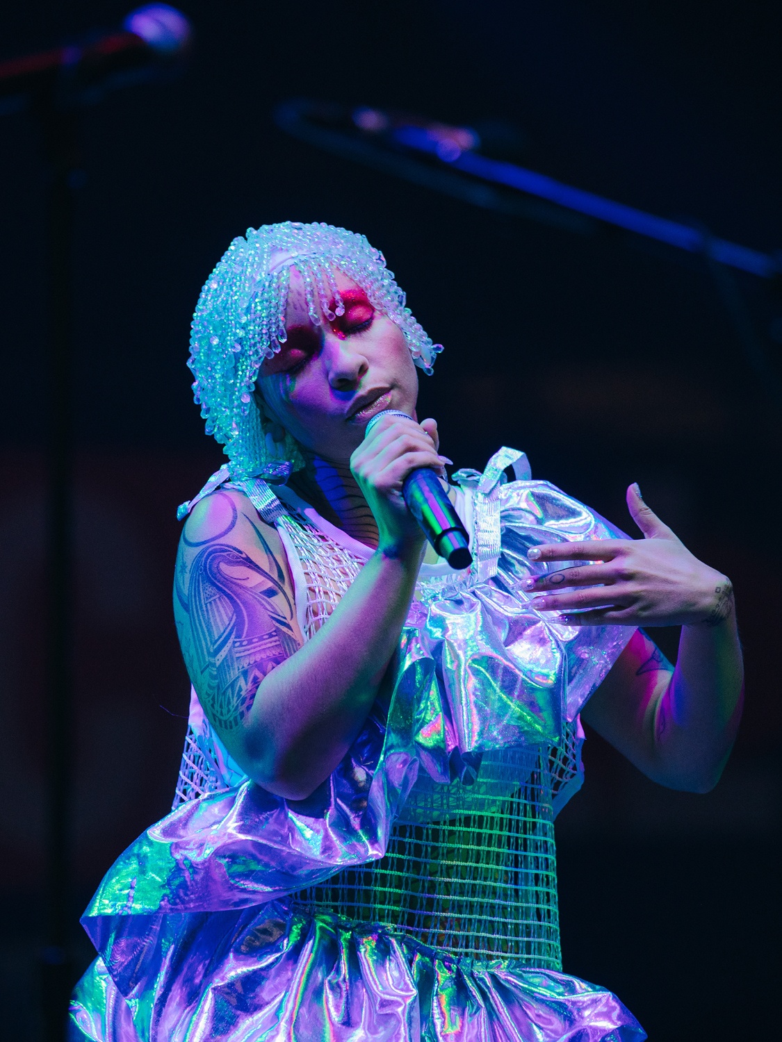 A performer sings into a microphone bathed in a bluish light. She wears a shimmering, beaded cap on her head and a shimmering ruffled costume.