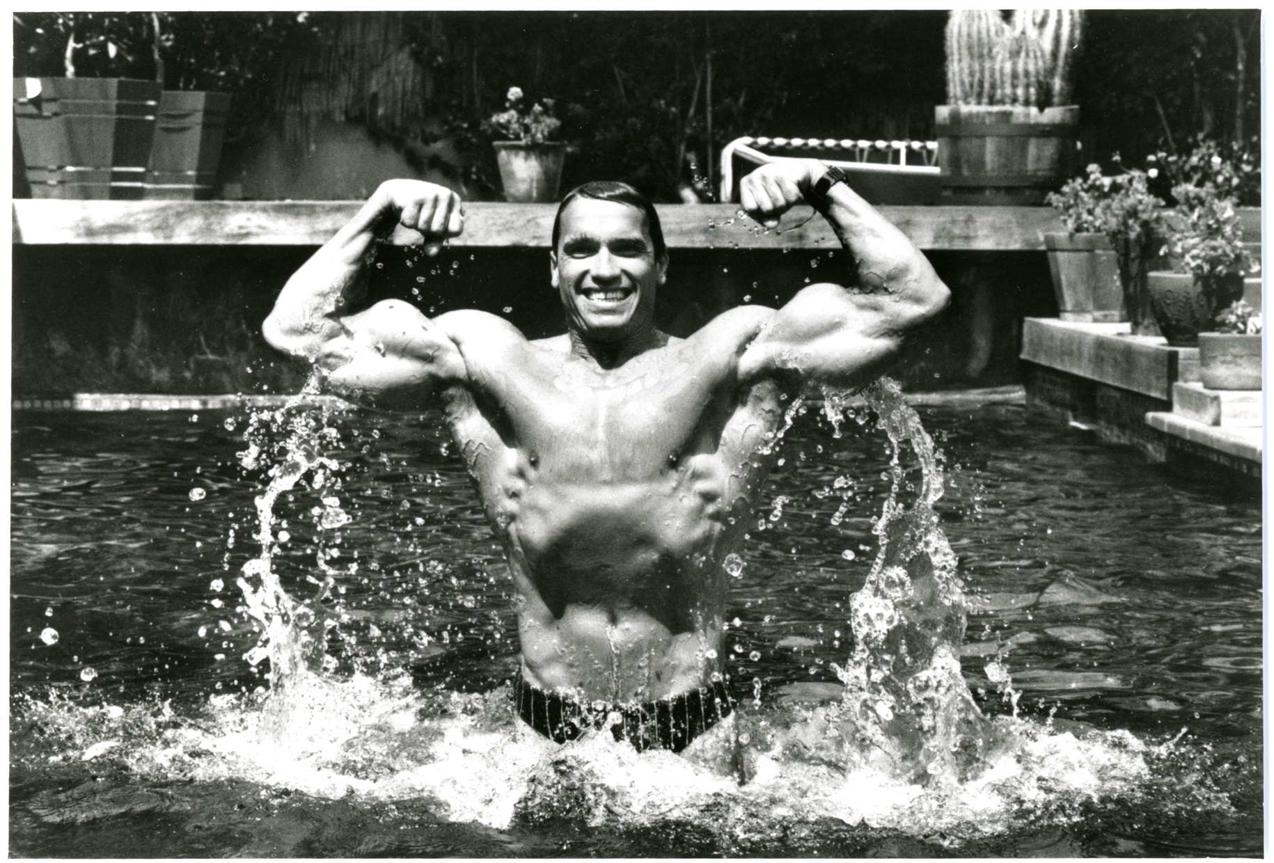 A black and white photograph of Arnold Schwarzenegger jumping out of a pool smiling and flexing.