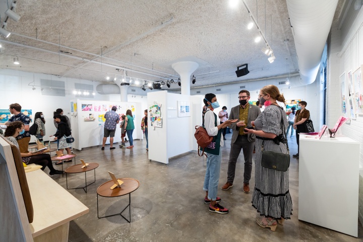 Exhibition view of wall dividers with posters on them at High Low. In the foreground three IVC faculty are in discussion.