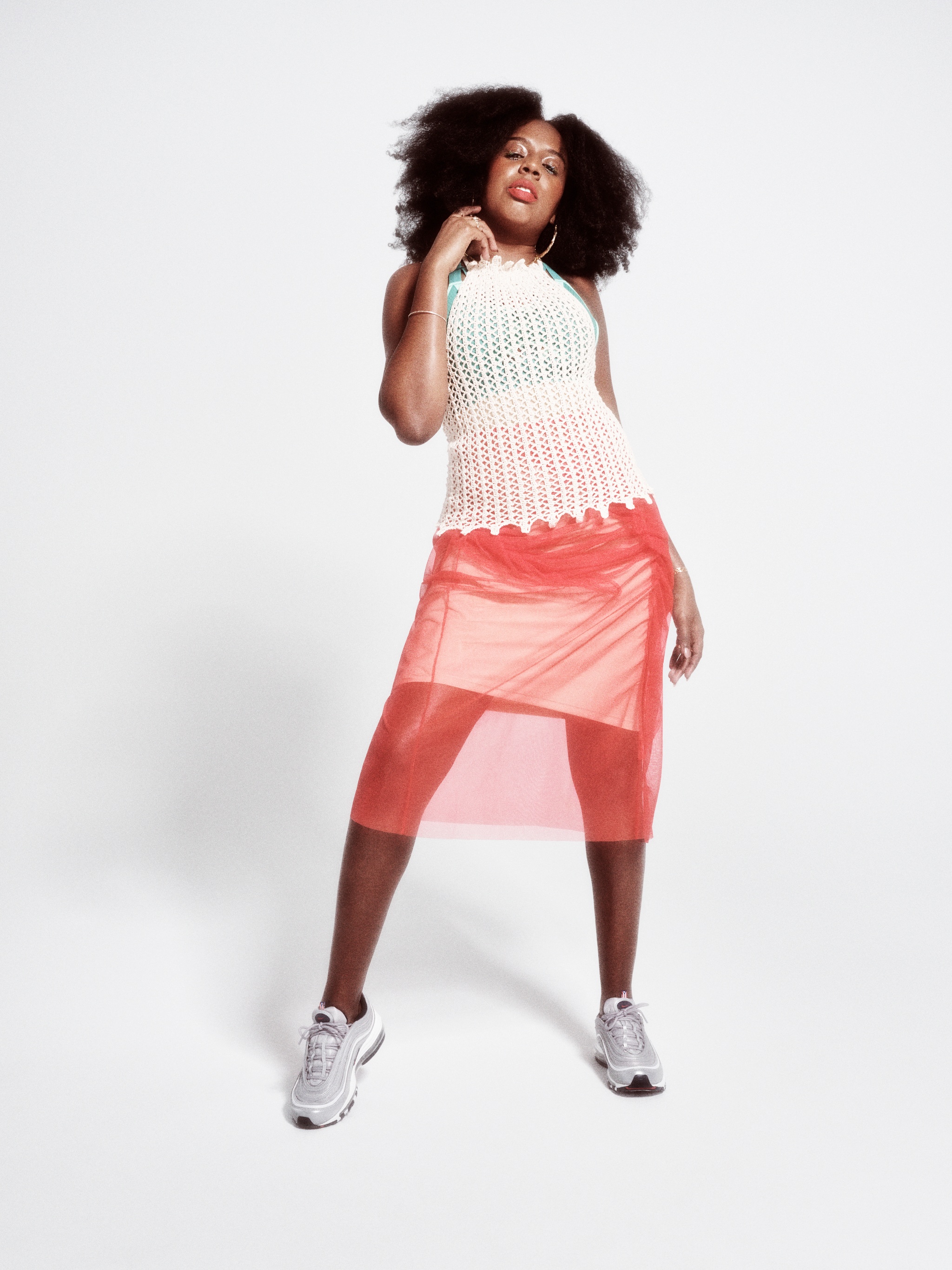 A portrait of DJ Bembona, a Black woman wearing a sheer red-orange skirt over a white dress. She wears a pair of Nike sneakers and stands against a white background.