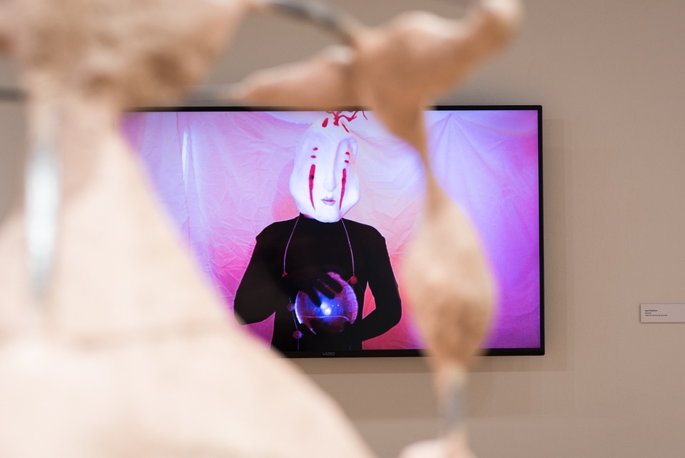 TV seen through a beige plaster sculpture. The tv displays a person in a white bug-like mask on a pink draped background holding an orb.