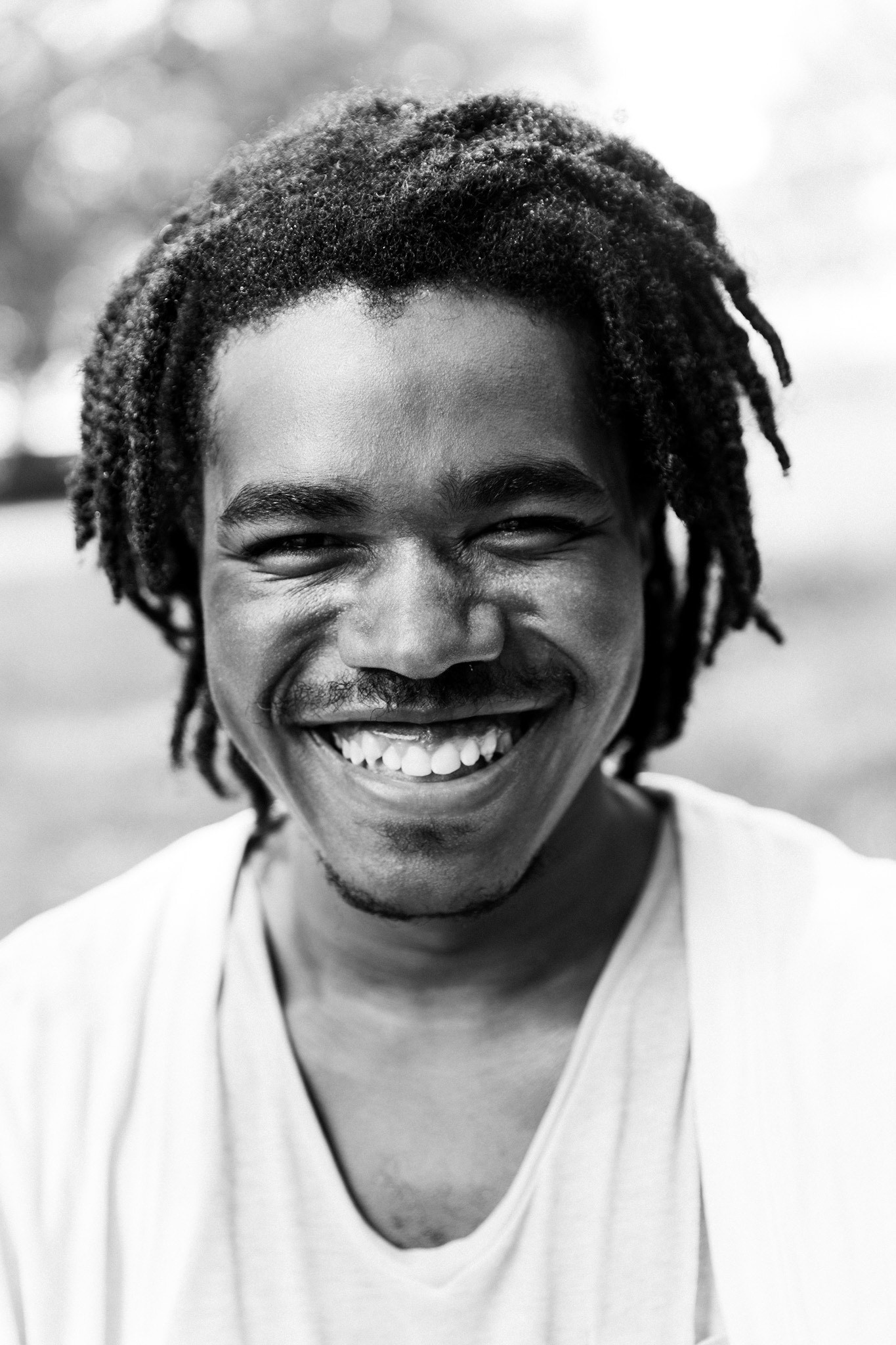 A portrait of a smiling Black man wearing a white v-neck t-shirt. He has a mustache across his upper lip and a beard that lines the area just under his chin. 