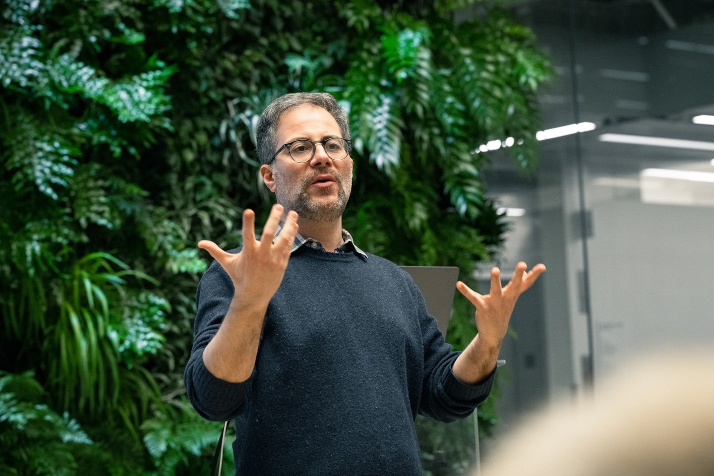 A man talks to an audience with a green plant wall behind him.