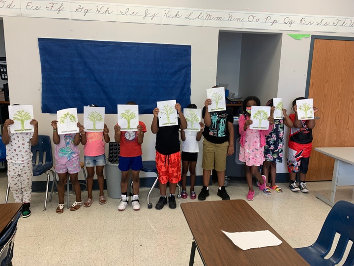 A row of young people hold up images of drawings of plants in a classroom. 