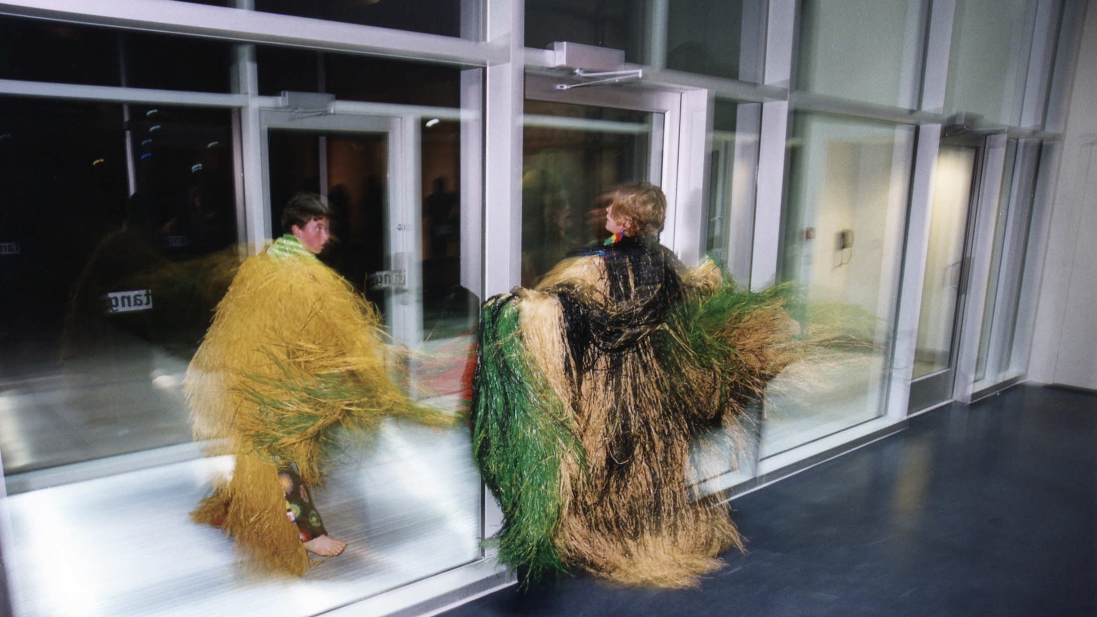 Two light-skinned people twirl in robes made of long grasses next to a glass entryway. 