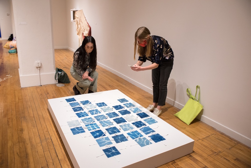 Two people bend over a short raised platform displaying a fabric work consisting of dozens of blue hand-dyed fabric squares the size of coasters, connected by short white fabric ties. 