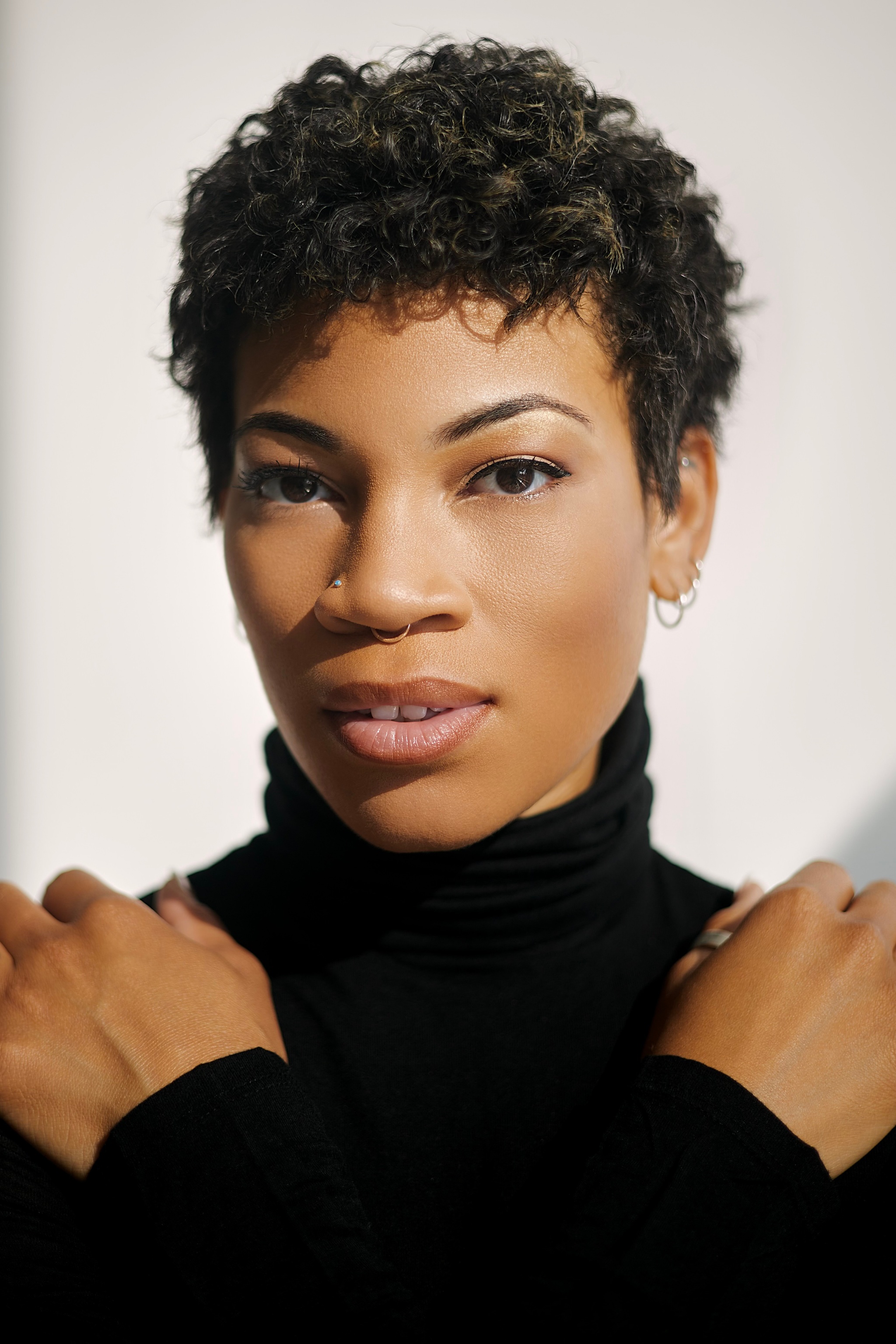 A woman with short hair looks into the camera and poses with her hands crossed over her chest so they rest on her shoulders. She wears a black turtleneck, and the background behind her is blank. 