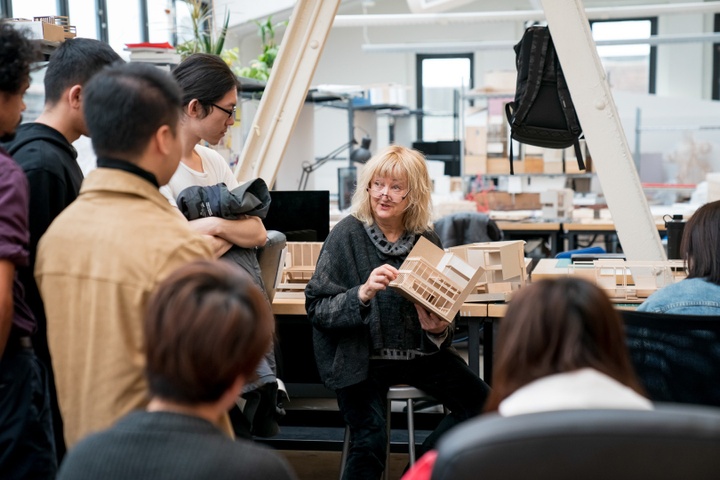Architect Yvonne Farrell sits in an architecture studio space in Givens Hall, holding a student's model and speaking to a group of students gathered around her.