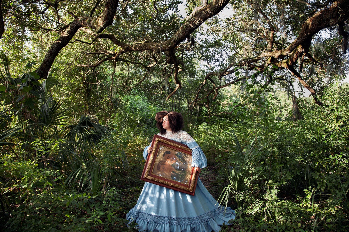 A woman carries a framed double-portrait an a verdant landscape