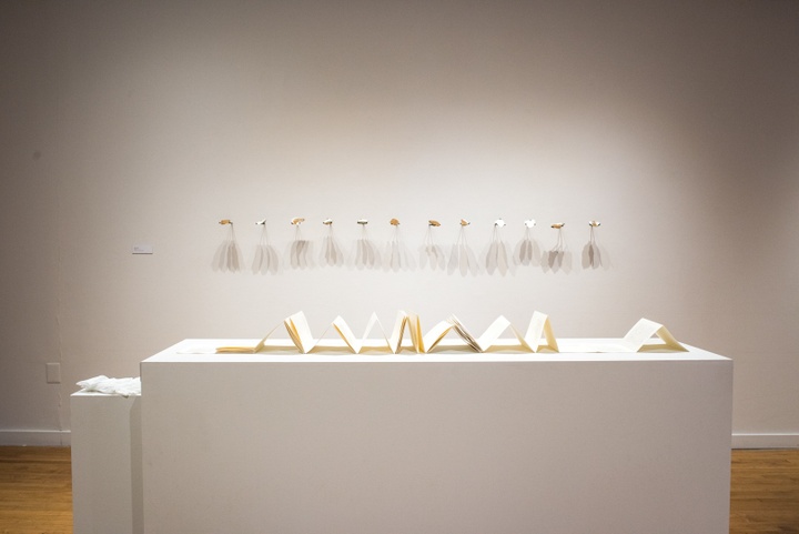 Wide shot of a book arts project on a display table. On the wall behind it is arrayed a row of oyster shells on forks.