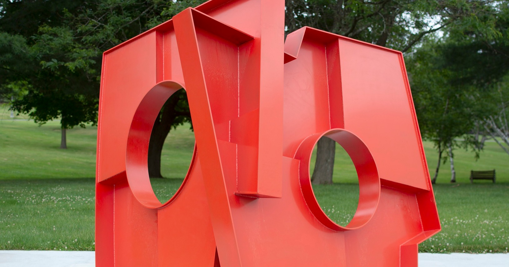 A large red sculpture of two rectangular shapes conjoined at an angle with large holes in the middle of each plane sits on a concrete slab in a grassy field with trees.
