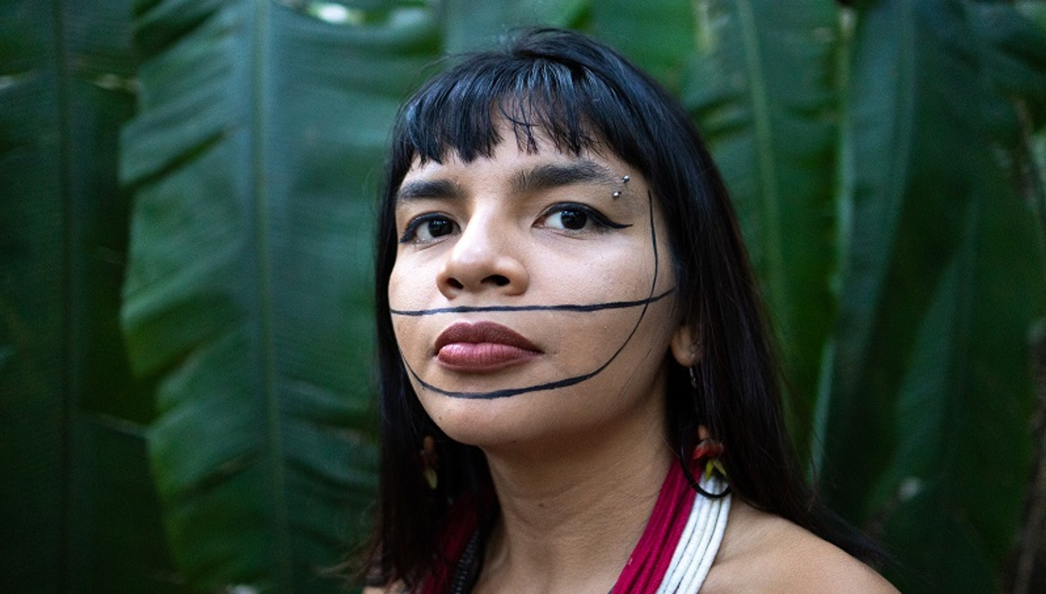 A portrait of Txai Suruí, and Indigenous woman from Brazil, who poses in front of large green leaves. She looks to the side and has a design of two black lines painted across her face. She has long black hair that falls beneath her shoulders and wears an eyebrow piercing at the end of her left eyebrow. 
