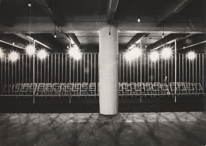 A black and white, landscape-oriented image from 1972 documenting Kate Millett’s installation called *Terminal Piece*. A barrier of wooden-slatted bars spans the length of an otherwise vacuous, cement industrial space. Illuminated from above, and set behind these bars, are two long rows of wooden folding chairs facing out towards the viewer. A mannequin of a lone, Caucasian, female-presenting figure is seated in one chair in the back row (located at the viewer’s right). The rest remain empty.