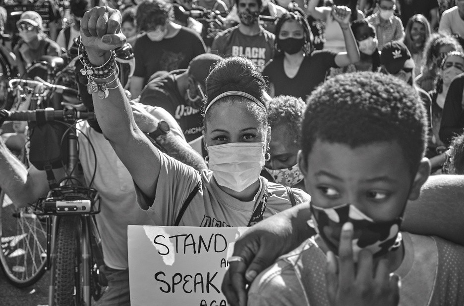 A black and white photograph of a crowd of protestors centered around a young, dark-skinned female wearing a mask, holding a sign and raising her right fist in the air.