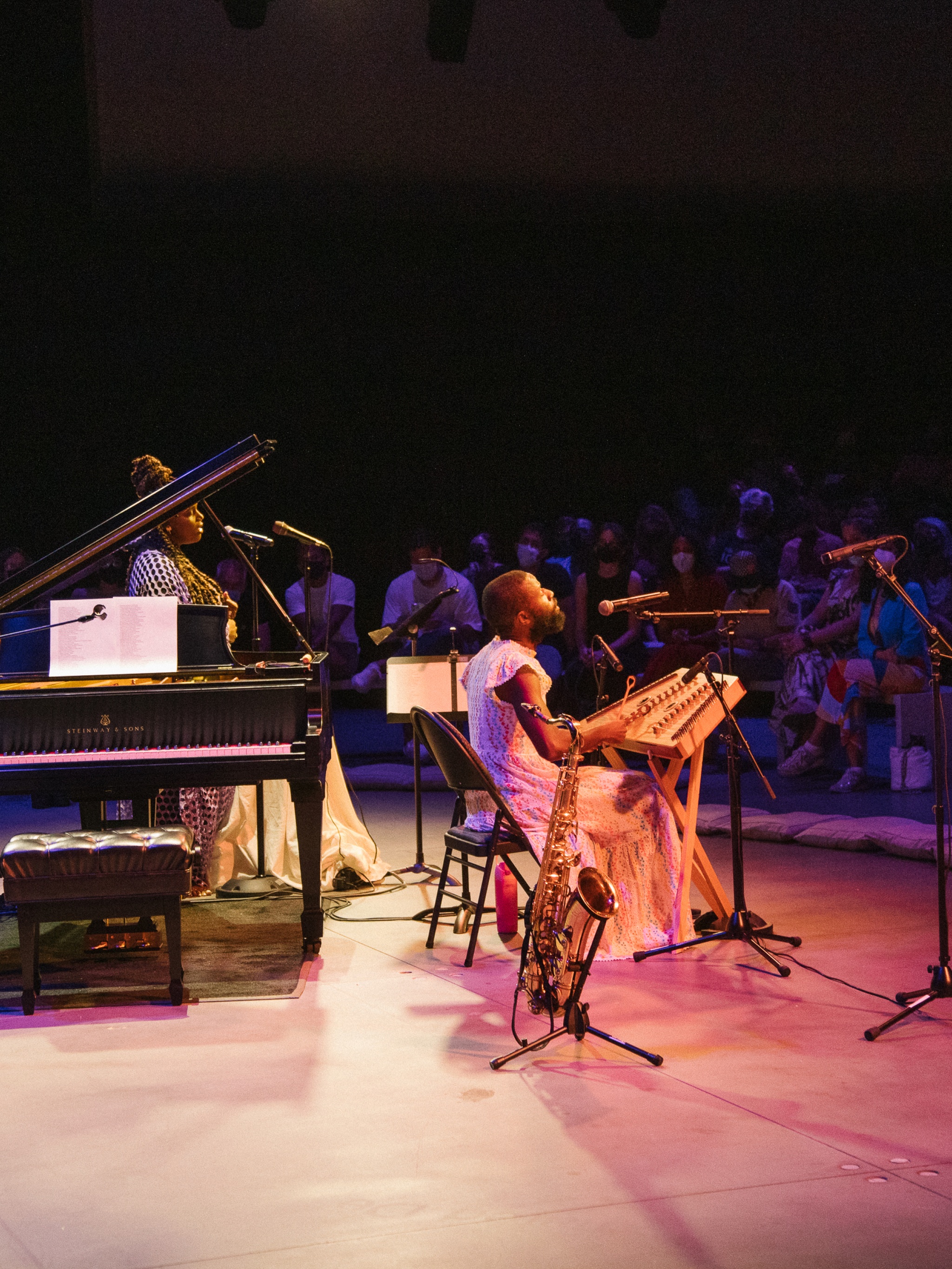The artist Jerome Ellis sits in front of an audience arranged in a semicircle in front of him in a darkened theater space. Jerome is a Black man who wears a long dress and has a beard. Beside him is a baby grand piano and a Black woman who is a singer.