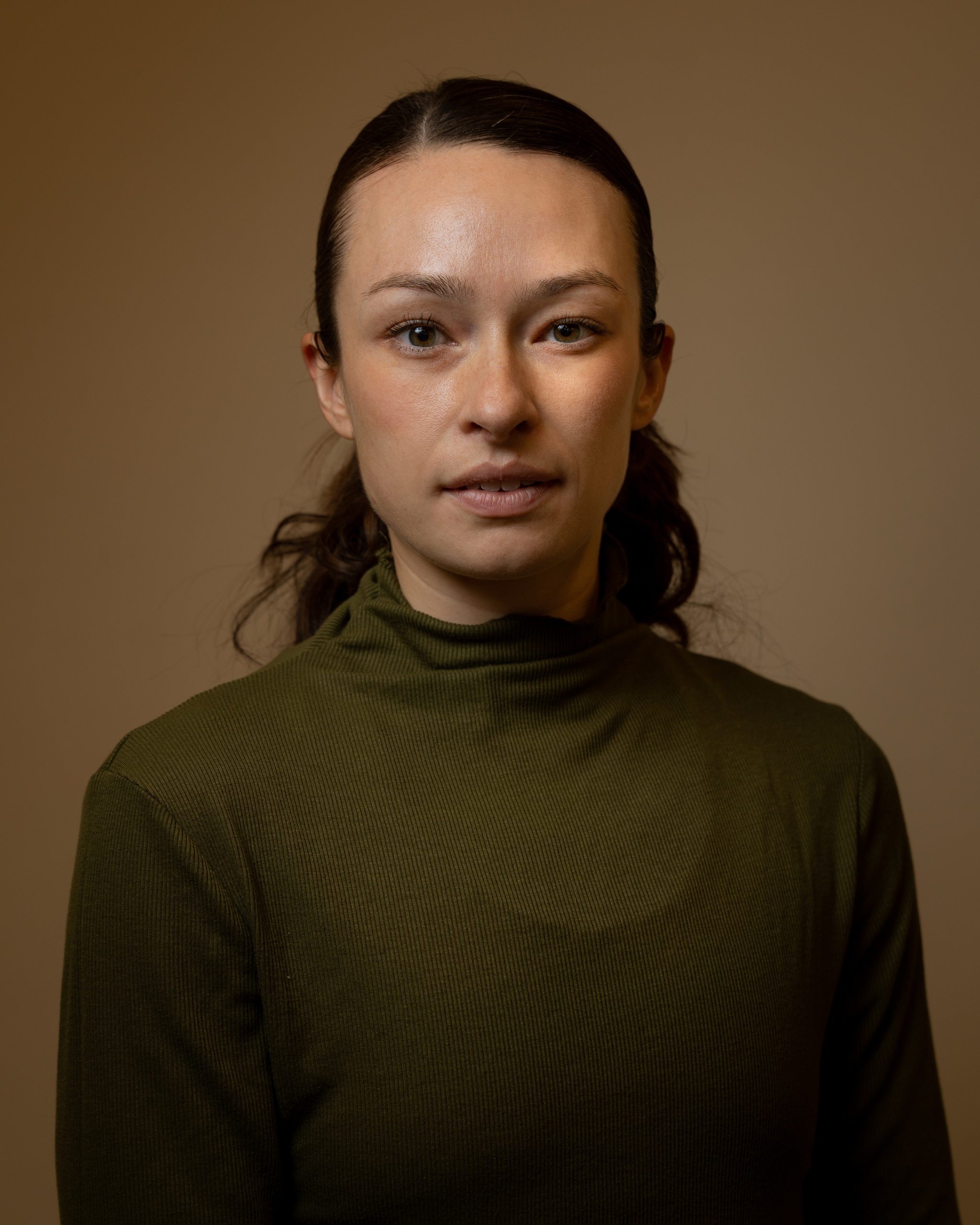A portrait of Nicole Baker against a neutral brown background. Nicole wears a dark, khaki green longsleeved shirt with her brown hair tied back behind her head. She looks directly at us with one eyebrow raised. 