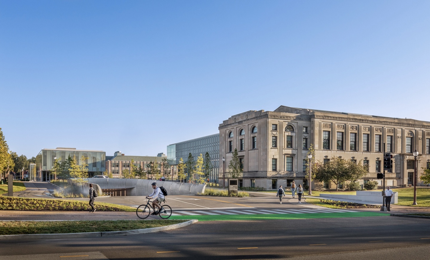 View from the street looking in at WashU's Danforth Campus. A paved drive leads into a covered underground parking structure. A Beaux-Arts-era building, Givens Hall, is prominent in the foreground to the right, with more two more modern buildings with glazed facades in the background. Pedestrians can be seen, including a person biking on a marked path.