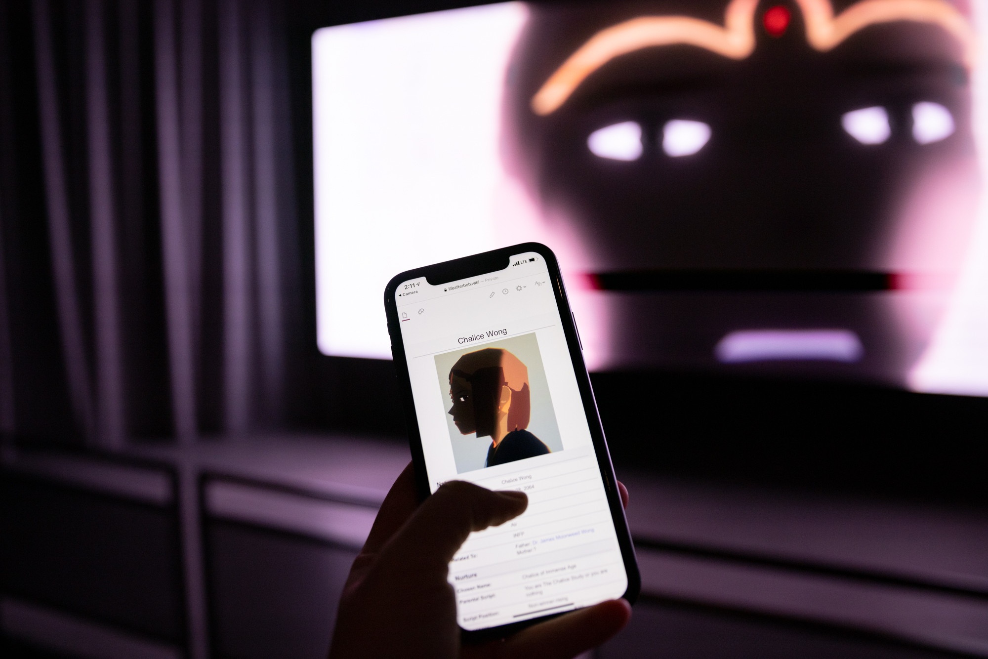 A hand holds a phone screen displaying a wiki page up in front of a movie screen with a close up of a character in an anime film.