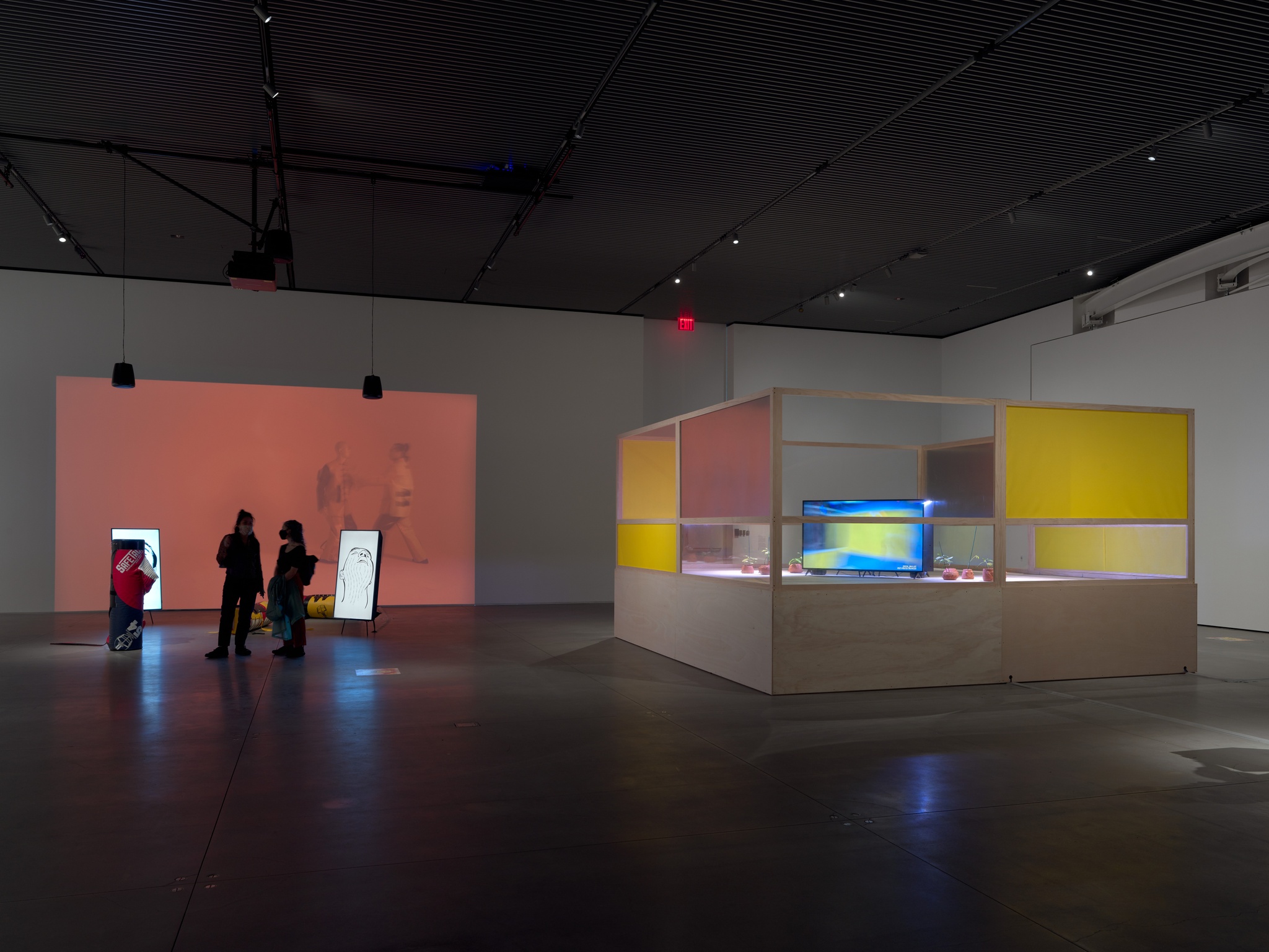 An art gallery with an installation resembling a boxing ring on the right and a video projection on the wall to the left. A couple people stand in front of the video projection.