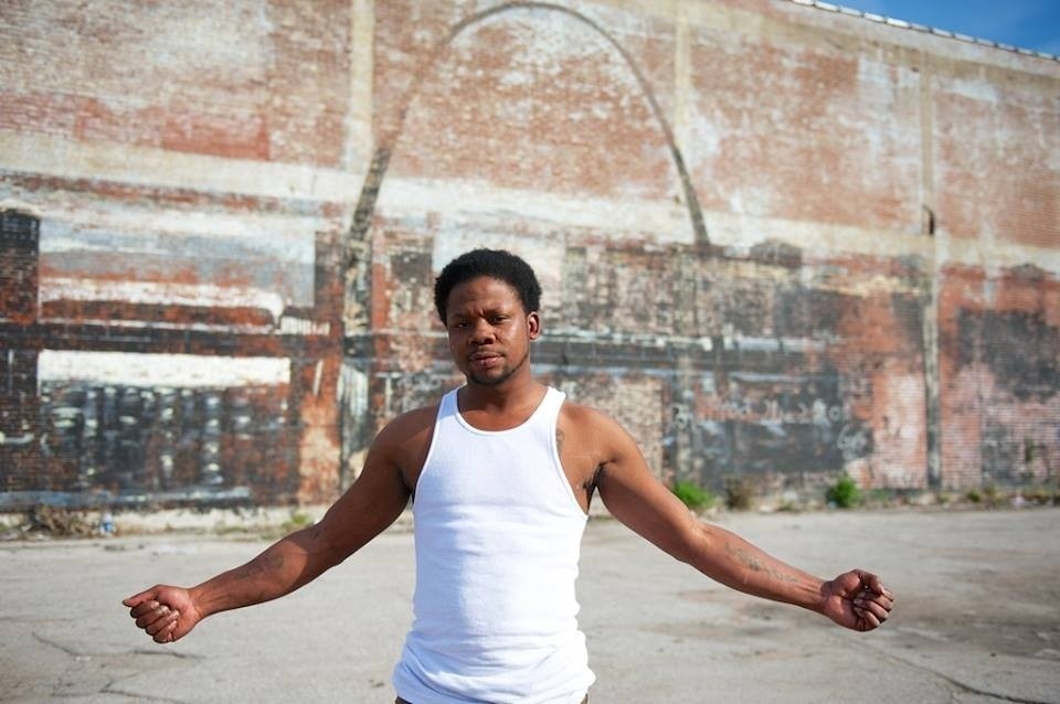 A man dressed in a white undershirt stands in front of a mural of the St. Louis skyline