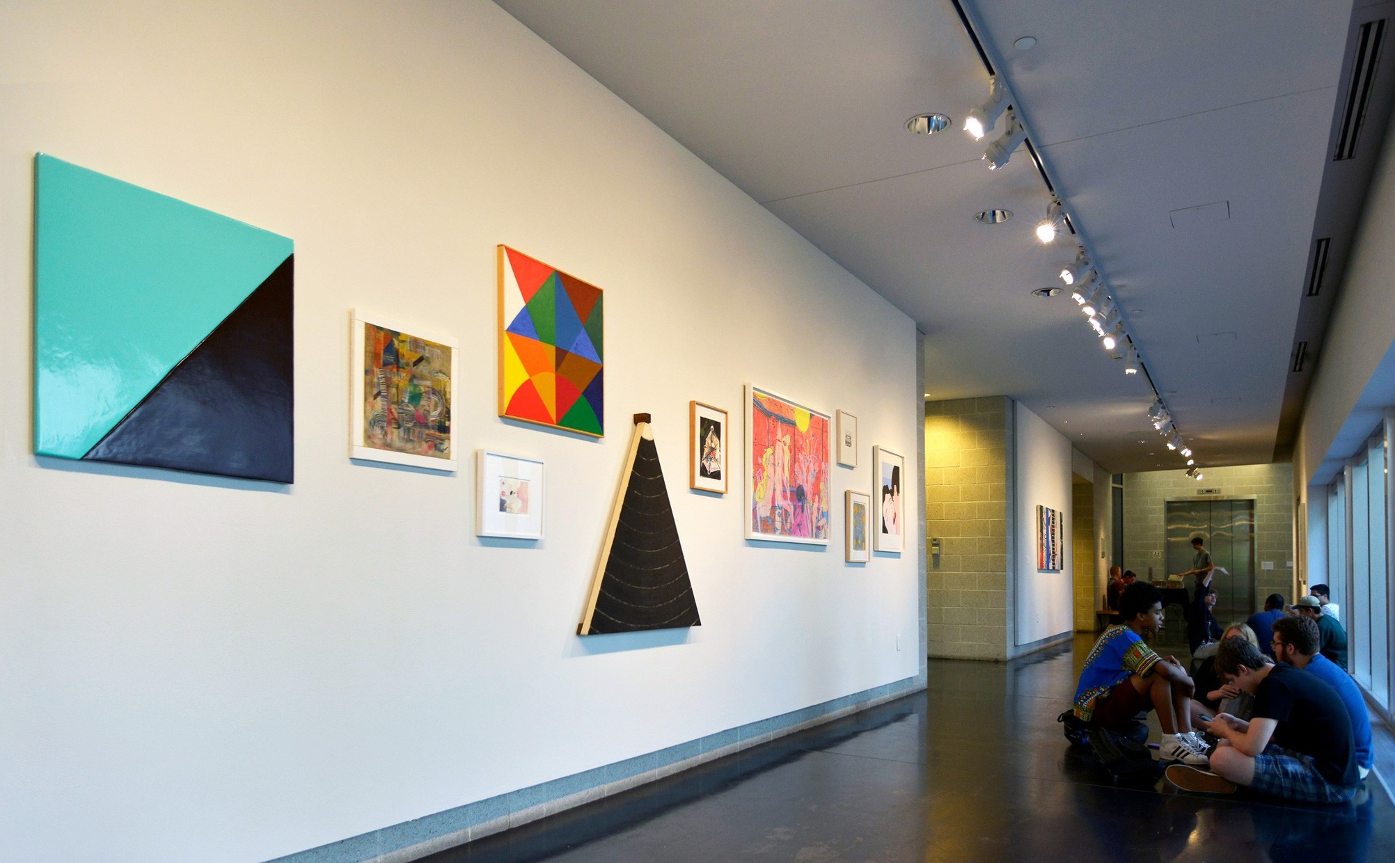 Colorful, abstract artworks hang on a white wall in a hallway. A group of young people sit on the floor opposite the wall of artwork.