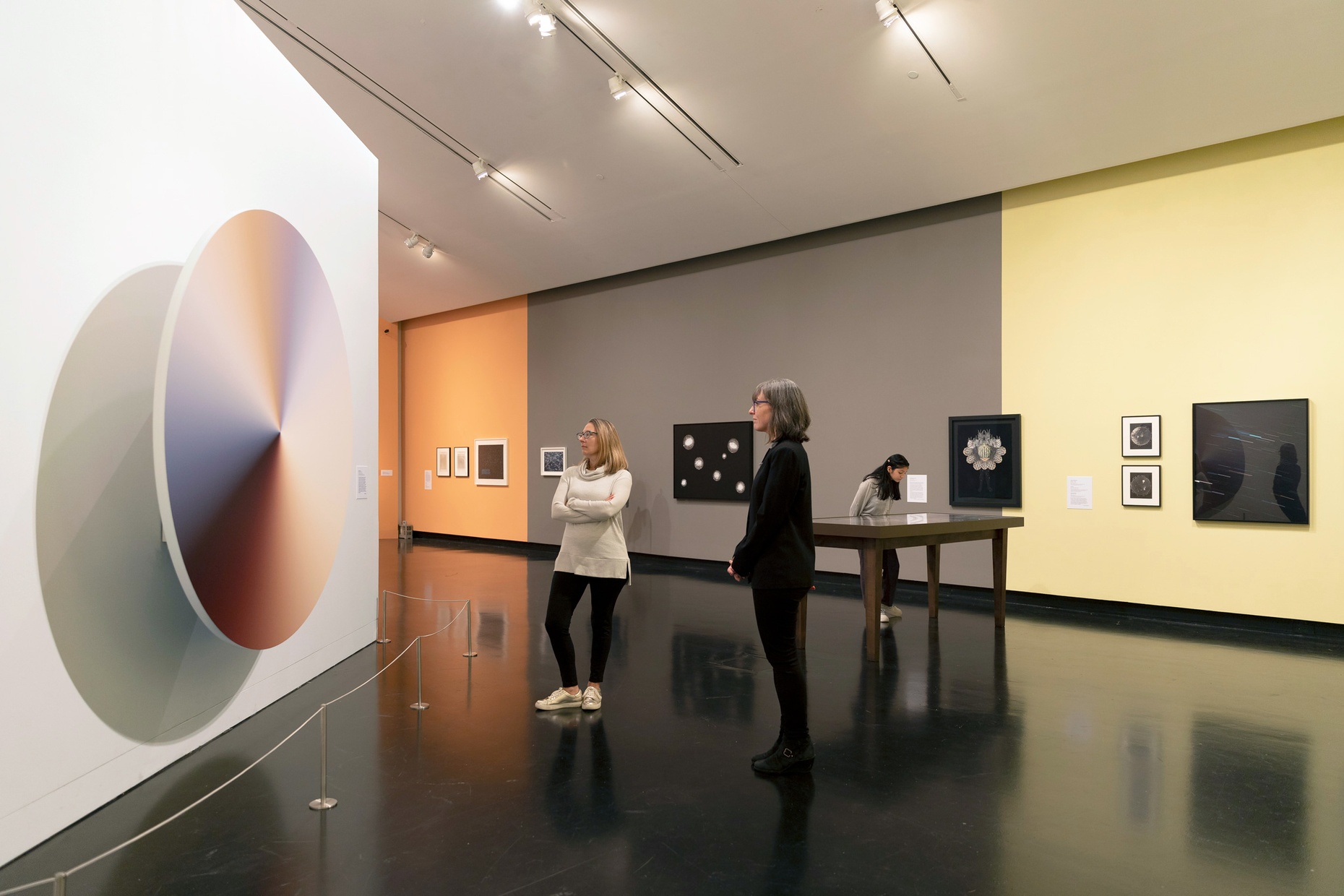 Two women stand and look to our left at a circular artwork on a white wall, diagonally in front of an orange, gray, and yellow wall with framed artworks.