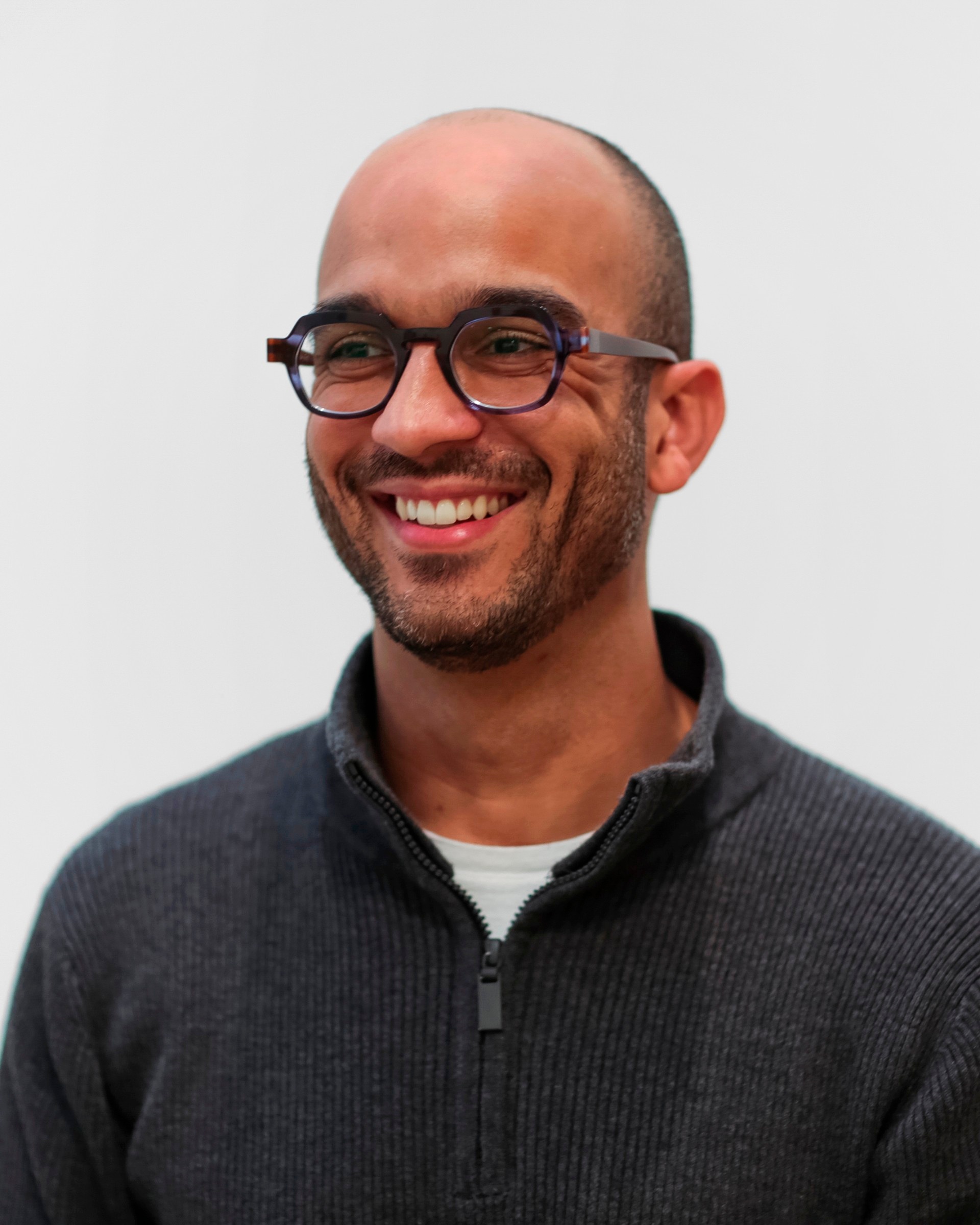 A photo of the artist Samuel Levi Jones against a white background. Jones is smiling, wearing glasses and a gray half-zip sweater.
