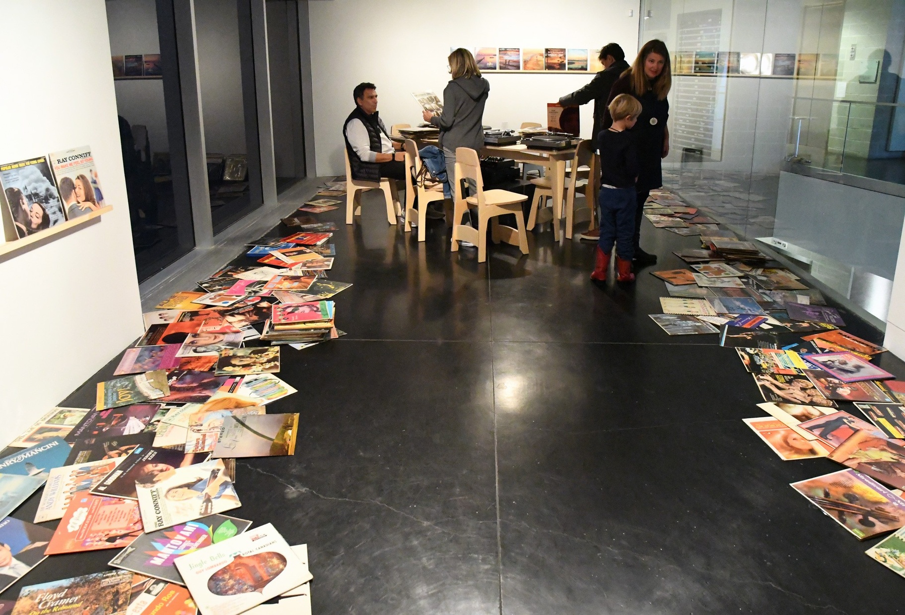 A group of light-skinned people gather around a wooden table with wooden chairs in a narrow room. Rows of vinyls hang on the walls and vinyls lay scattered on the floor around the edges of the room.