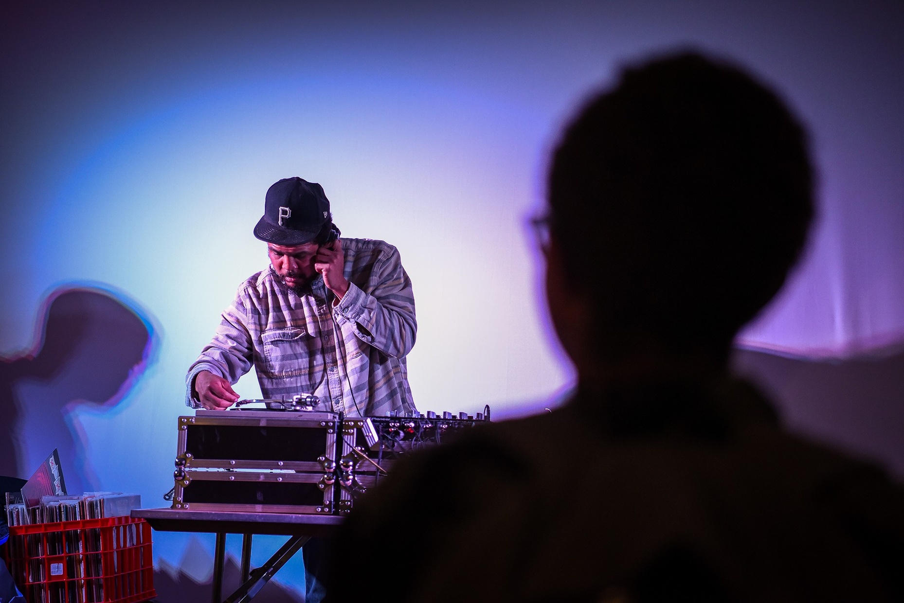 Photo of a man deejaying holding headphones to his ear with the silhouette of a woman in the foreground.