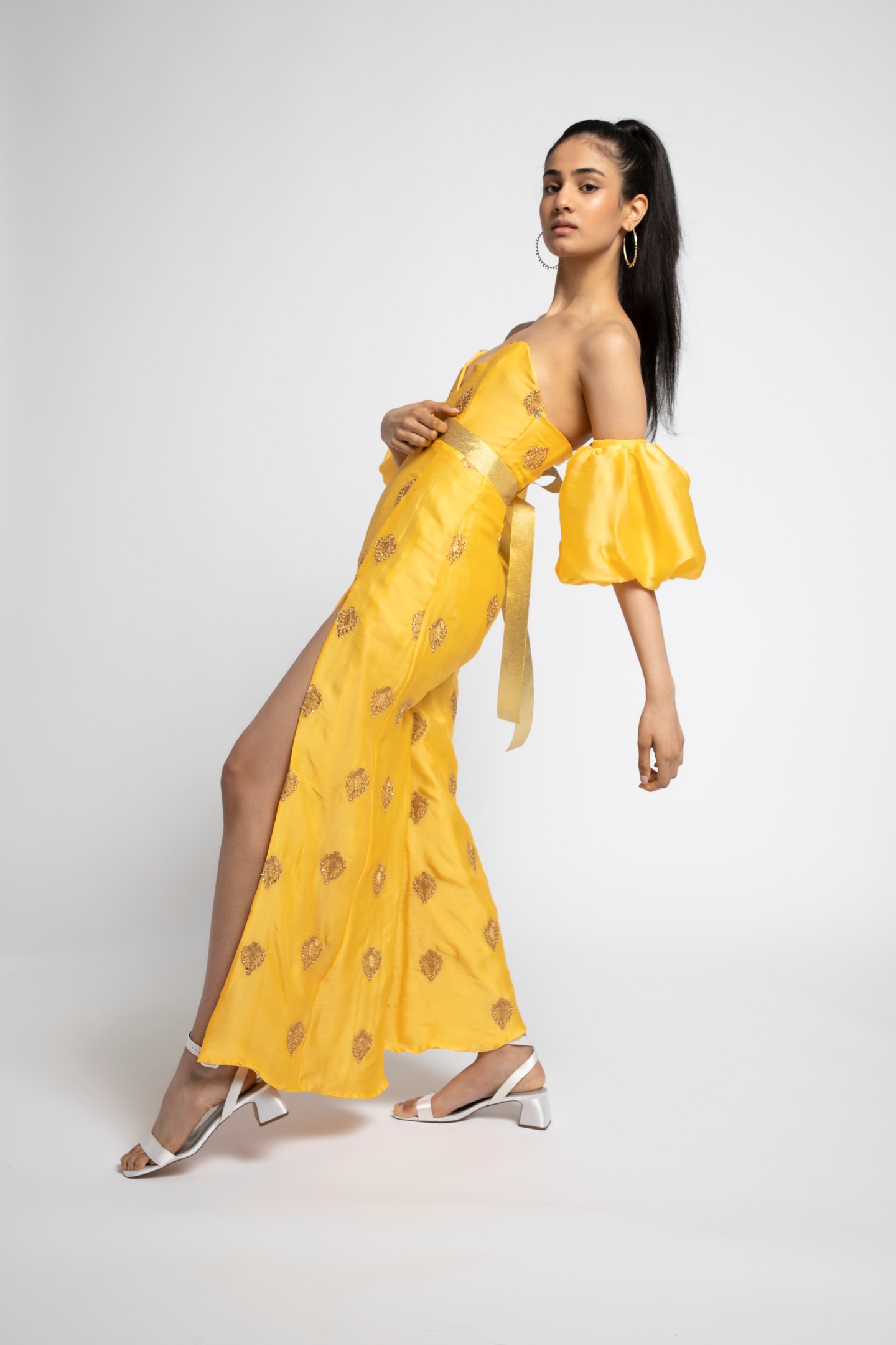 Model in a yellow strapless jumpsuit made of bright yellow fabric with a pattern of golden diamond-shaped motifs and cinched by a gold ribbon waistband. The jumpsuit legs are slit to the upper thigh on the front creating a skirtlike sweep. The model wears two detached sleeve puffs on her upper arms.