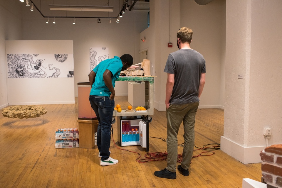 Two people in a gallery space peer at a construction made from a minifridge, a cart, an endtable, and an aquarium tank full of murky orange liquid.