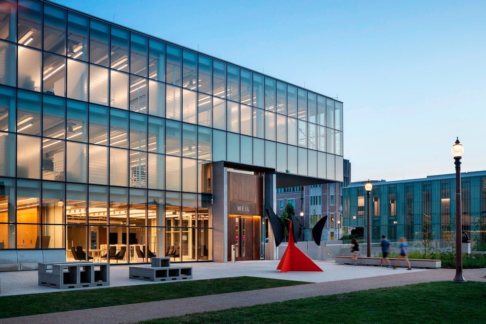 Exterior photo of the three-story, contemporary building Weil Hall, with three levels of windows lit up at dusk. Additional campus buildings can be seen in the background; an orange Alexander Calder sculpture is visible in the foreground.